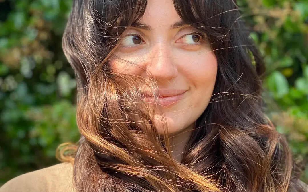 Smiling woman outdoors with long dark hair featuring a warm caramel balayage and sweeping curtain bangs softly blowing in the wind, custom colored by Lisa Edwards at Sunday Salon in Raleigh, NC.