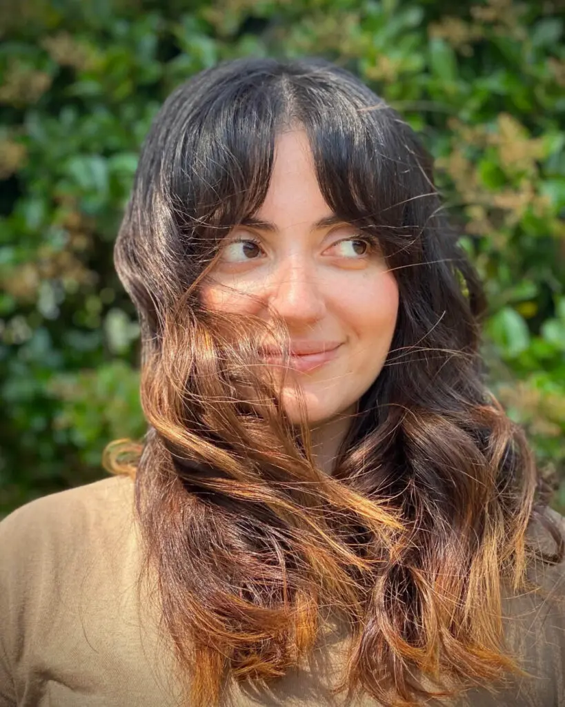 Sun-Kissed Warm Caramel Balayage with Effortless Curtain Bangs Smiling woman outdoors with long dark hair featuring a warm caramel balayage and sweeping curtain bangs softly blowing in the wind, custom colored by Lisa Edwards at Sunday Salon in Raleigh, NC.