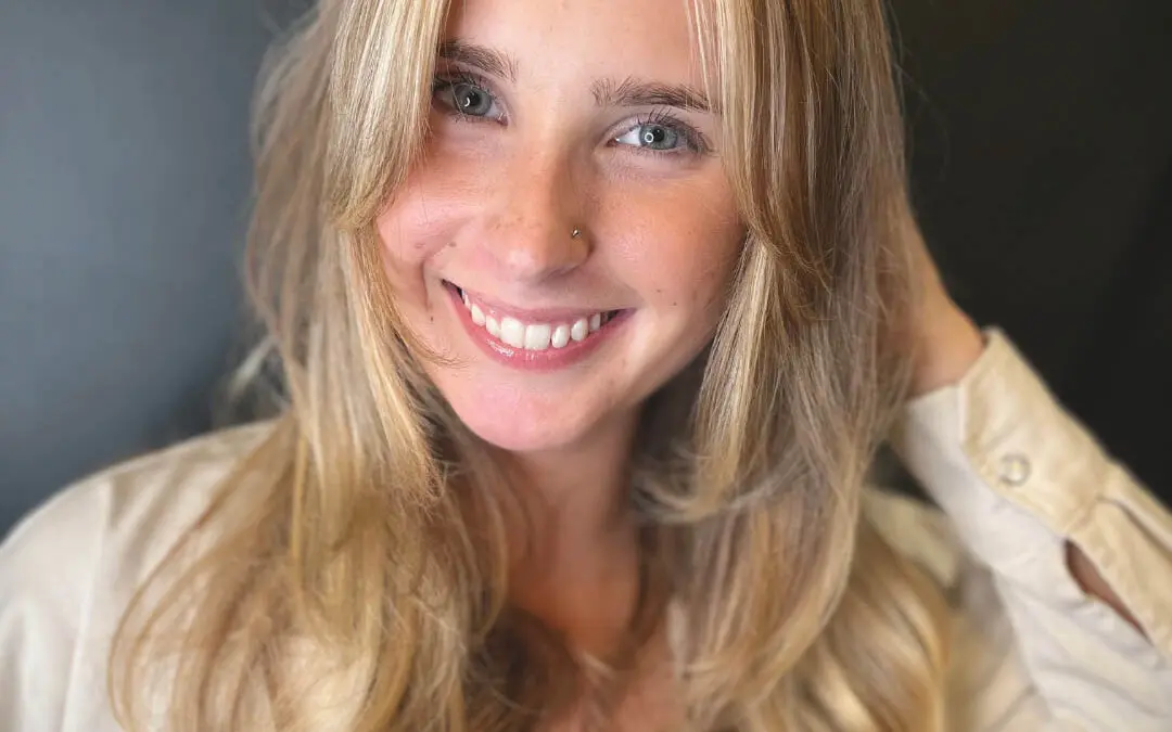 Close-up photo of a smiling woman with a voluminous blonde butterfly haircut and curtain bangs styled in a bouncy blowout, created by Katherine Beingolea at Sunday Salon in Raleigh, NC.