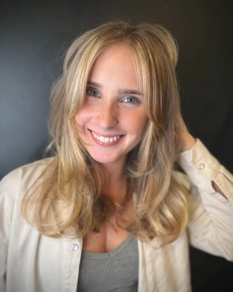 Close-up photo of a smiling woman with a voluminous blonde butterfly haircut and curtain bangs styled in a bouncy blowout, created by Katherine Beingolea at Sunday Salon in Raleigh, NC.