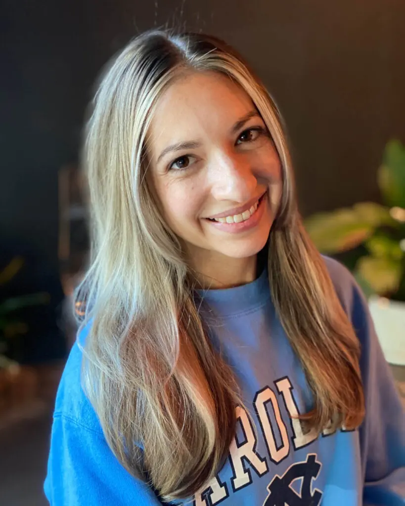 Close-up photo of a smiling client showcasing long sleek bronde hair with bright blonde face-framing highlights and a shadow root, custom colored by Lisa Edwards at Sunday Salon in Raleigh, NC.