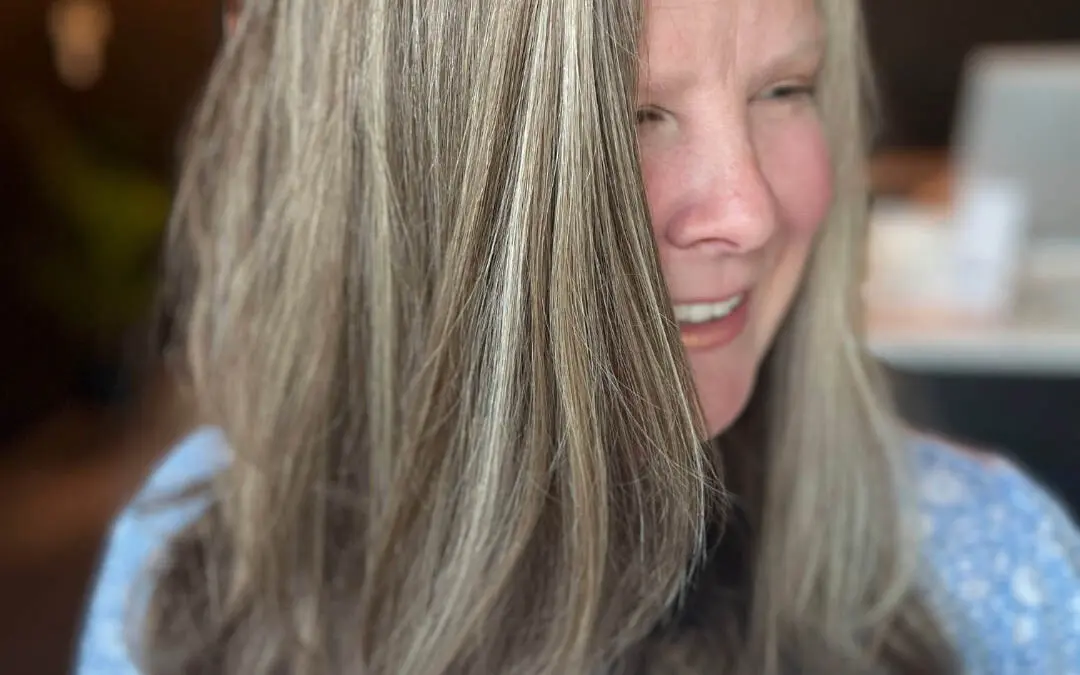 A smiling client showcasing medium-long hair with dimensional grey blending and silver-blonde highlights, styled with soft waves at the ends, custom colored by Lisa Edwards at Sunday Salon in Raleigh, NC.