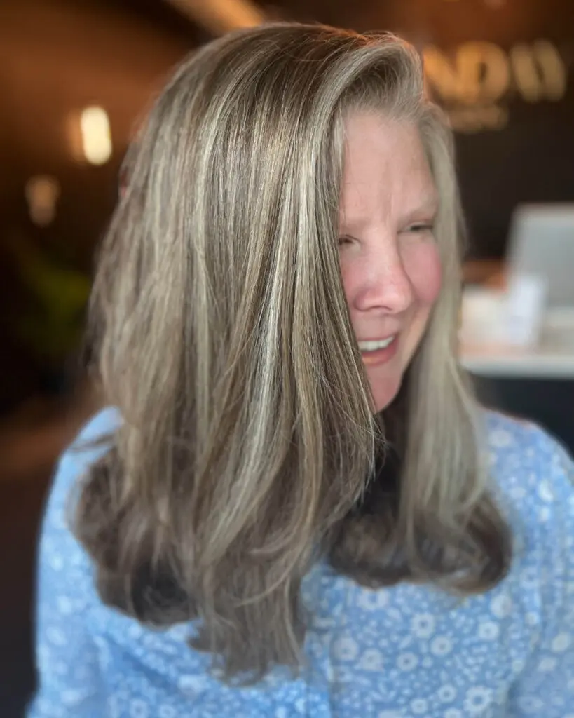 A smiling client showcasing medium-long hair with dimensional grey blending and silver-blonde highlights, styled with soft waves at the ends, custom colored by Lisa Edwards at Sunday Salon in Raleigh, NC.
