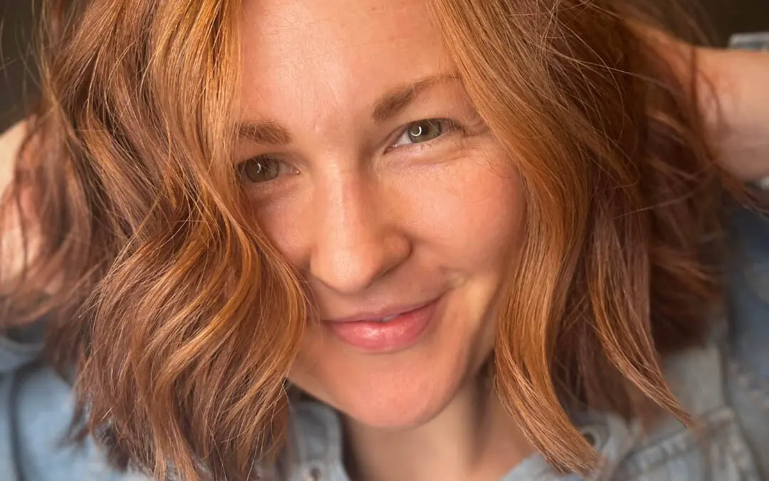 Close-up photo of a smiling client running hands through her vibrant copper hair styled in a textured bob with tousled waves, custom colored by Lisa Edwards at Sunday Salon in Raleigh, NC.