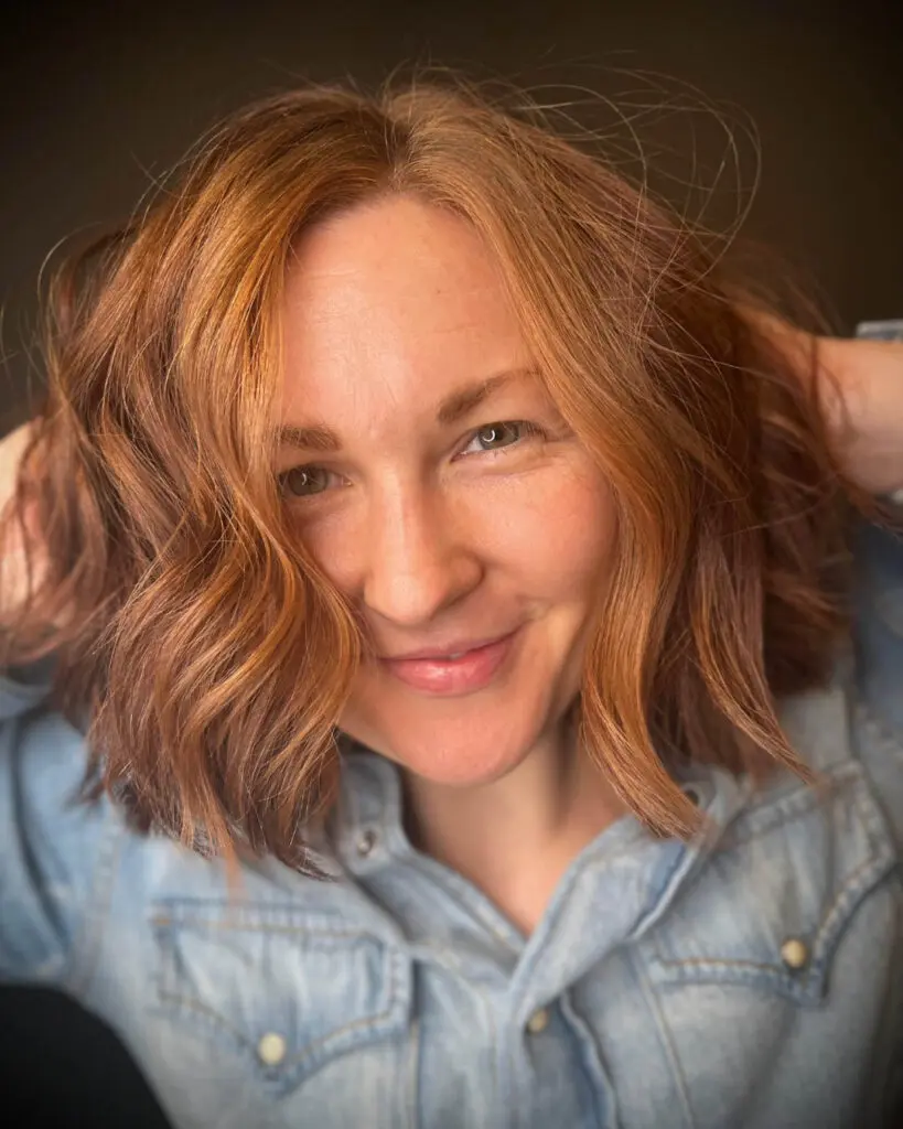 Close-up photo of a smiling client running hands through her vibrant copper hair styled in a textured bob with tousled waves, custom colored by Lisa Edwards at Sunday Salon in Raleigh, NC.