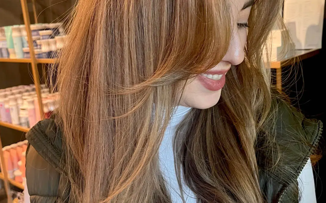 Side profile of a smiling client with long, warm caramel balayage hair featuring swooping face-framing layers, styled in a sleek blowout at Sunday Salon in Raleigh, NC.