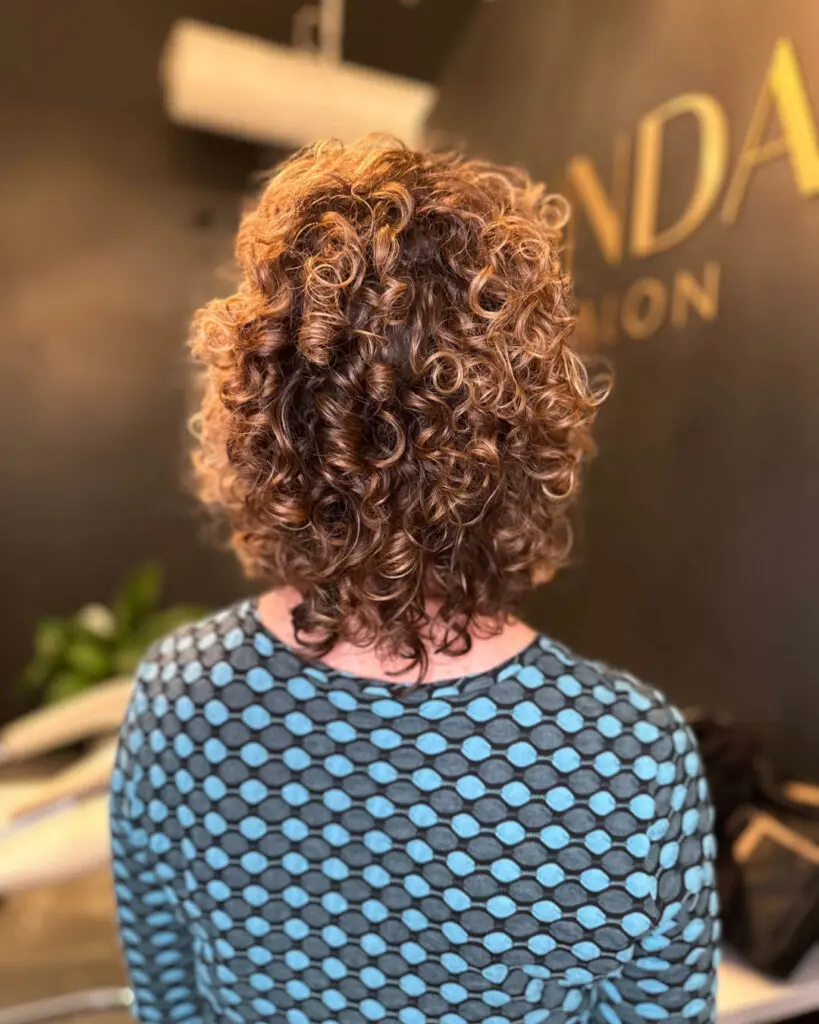 Dimensional Golden Caramel Highlights on Voluminous Natural Curls Back view of a client with defined, shoulder-length natural curls featuring dimensional golden caramel highlights, styled to perfection at Sunday Salon in Raleigh, NC.