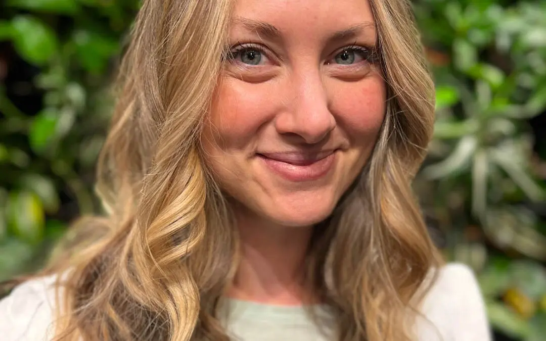 Smiling client with long warm honey blonde balayage hair and a subtle shadow root styled in loose cascading waves at Sunday Salon in Raleigh, NC.