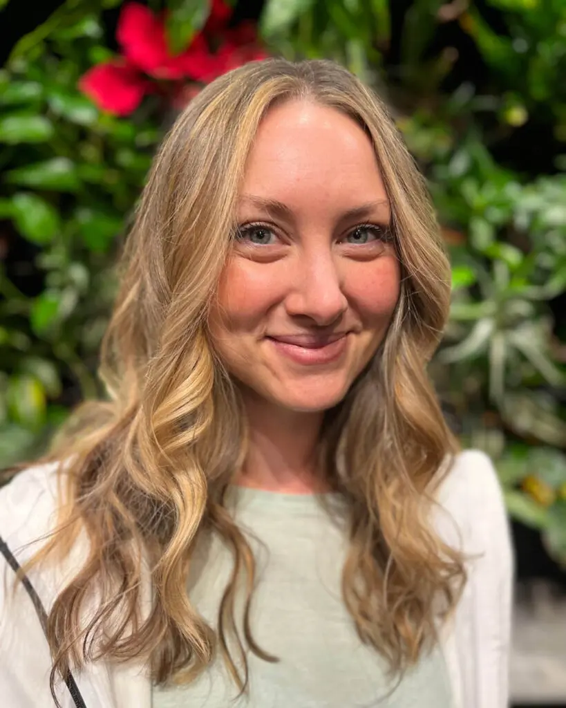 Smiling client with long warm honey blonde balayage hair and a subtle shadow root styled in loose cascading waves at Sunday Salon in Raleigh, NC.
