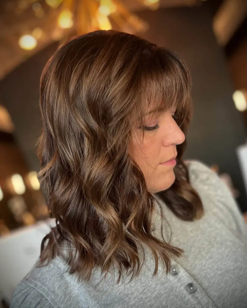 Dimensional Chocolate Brunette Lob with Wispy Bangs and Caramel Highlights Side profile of a client with a dimensional chocolate brunette lob, wispy bangs, and subtle caramel highlights styled in soft waves by Katherine Beingolea at Sunday Salon in Raleigh, NC.