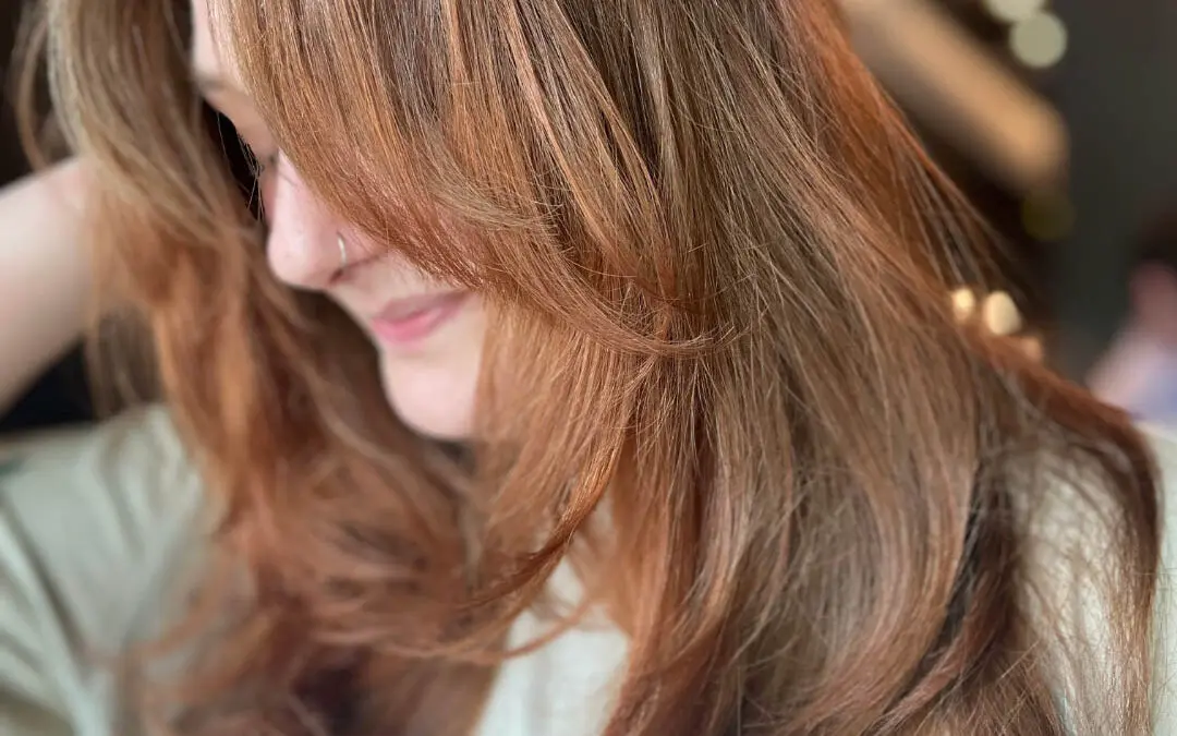Side profile of a smiling client with long warm auburn brunette hair and sweeping curtain bangs styled in a voluminous blowout at Sunday Salon in Raleigh, NC.