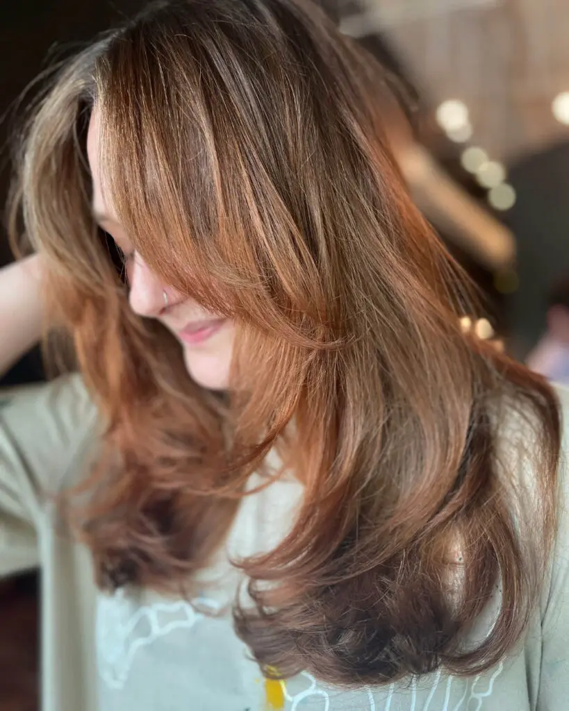 Voluminous Warm Auburn Brunette Blowout with Sweeping Curtain Bangs Side profile of a smiling client with long warm auburn brunette hair and sweeping curtain bangs styled in a voluminous blowout at Sunday Salon in Raleigh, NC.