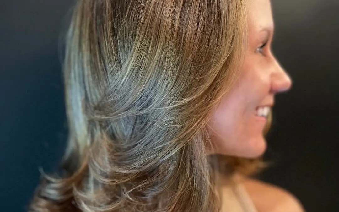 Side profile of a smiling client showcasing a dimensional golden bronde hair color with heavily layered, bouncy flipped ends styled in a smooth blowout at Sunday Salon in Raleigh, NC.