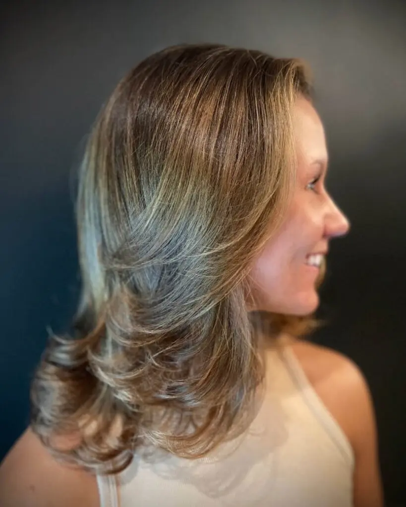 Side profile of a smiling client showcasing a dimensional golden bronde hair color with heavily layered, bouncy flipped ends styled in a smooth blowout at Sunday Salon in Raleigh, NC.