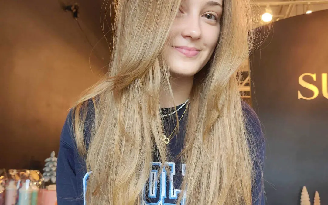 Smiling client showcasing ultra-long warm sandy blonde hair with a soft shadow root and sweeping face-framing layers styled in a smooth blowout at Sunday Salon in Raleigh, NC.