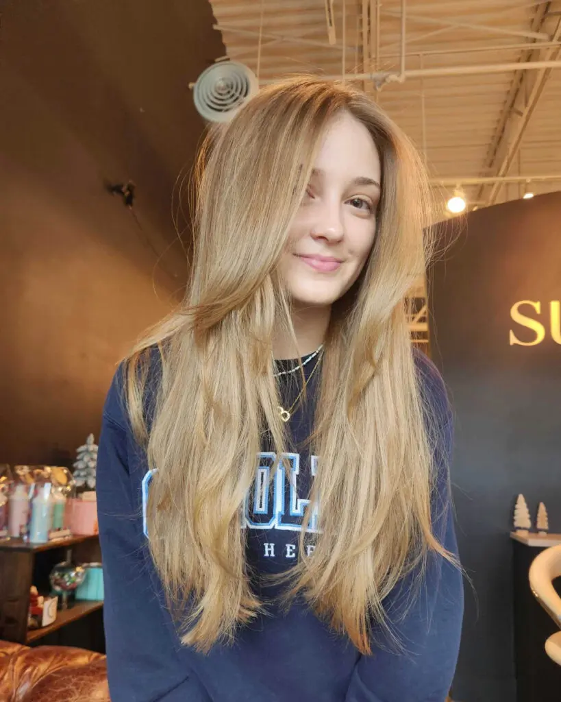 Smiling client showcasing ultra-long warm sandy blonde hair with a soft shadow root and sweeping face-framing layers styled in a smooth blowout at Sunday Salon in Raleigh, NC.