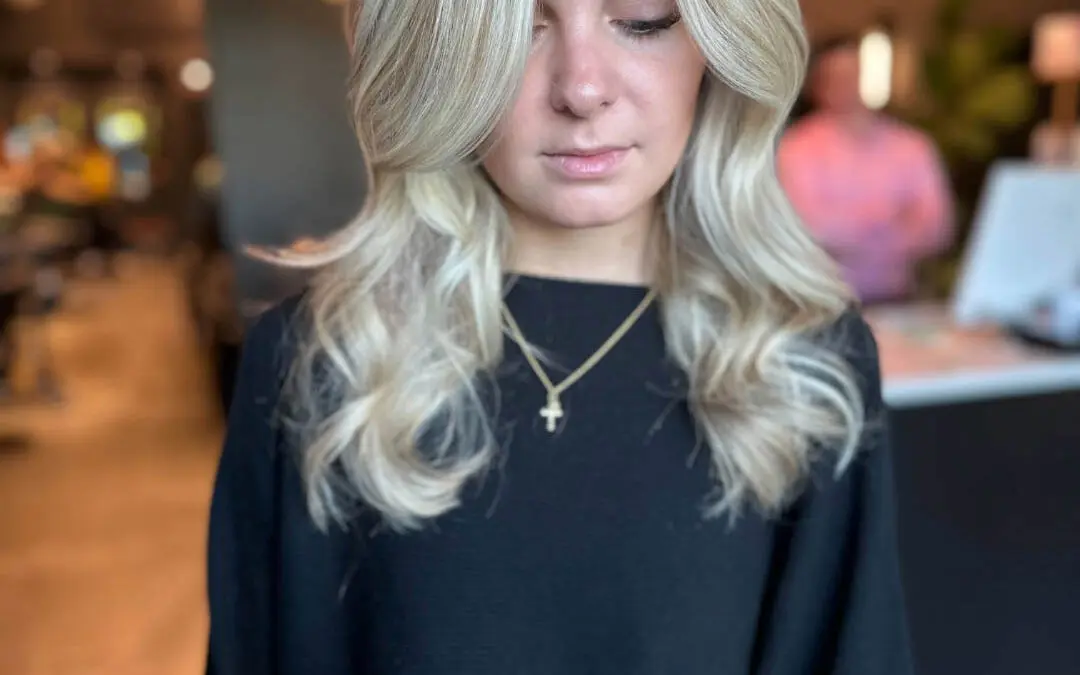 Woman looking down showcasing medium-long, bright icy vanilla blonde hair styled with a voluminous blowout and sweeping curtain bangs, custom colored by Lisa Edwards at Sunday Salon in Raleigh, NC.