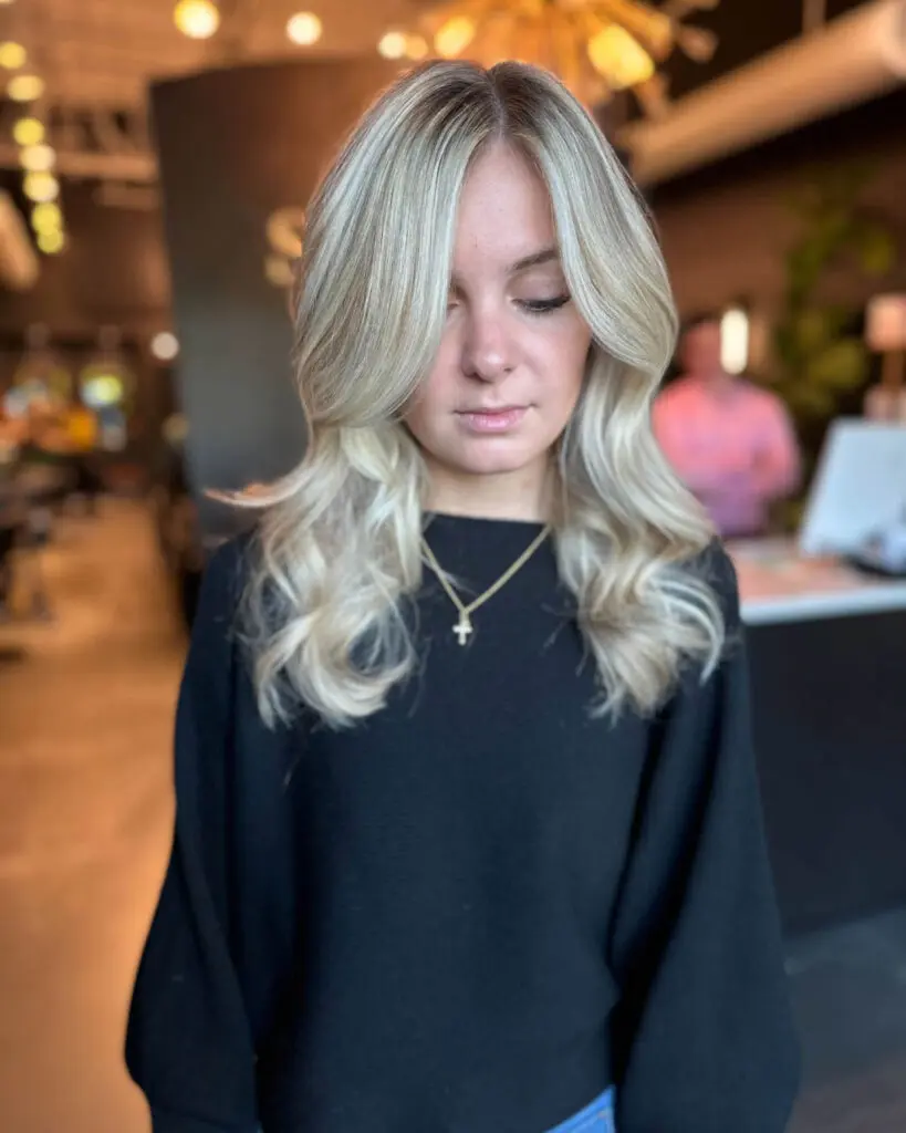Bright Icy Vanilla Blonde with Voluminous Curtain Bangs Woman looking down showcasing medium-long, bright icy vanilla blonde hair styled with a voluminous blowout and sweeping curtain bangs, custom colored by Lisa Edwards at Sunday Salon in Raleigh, NC.