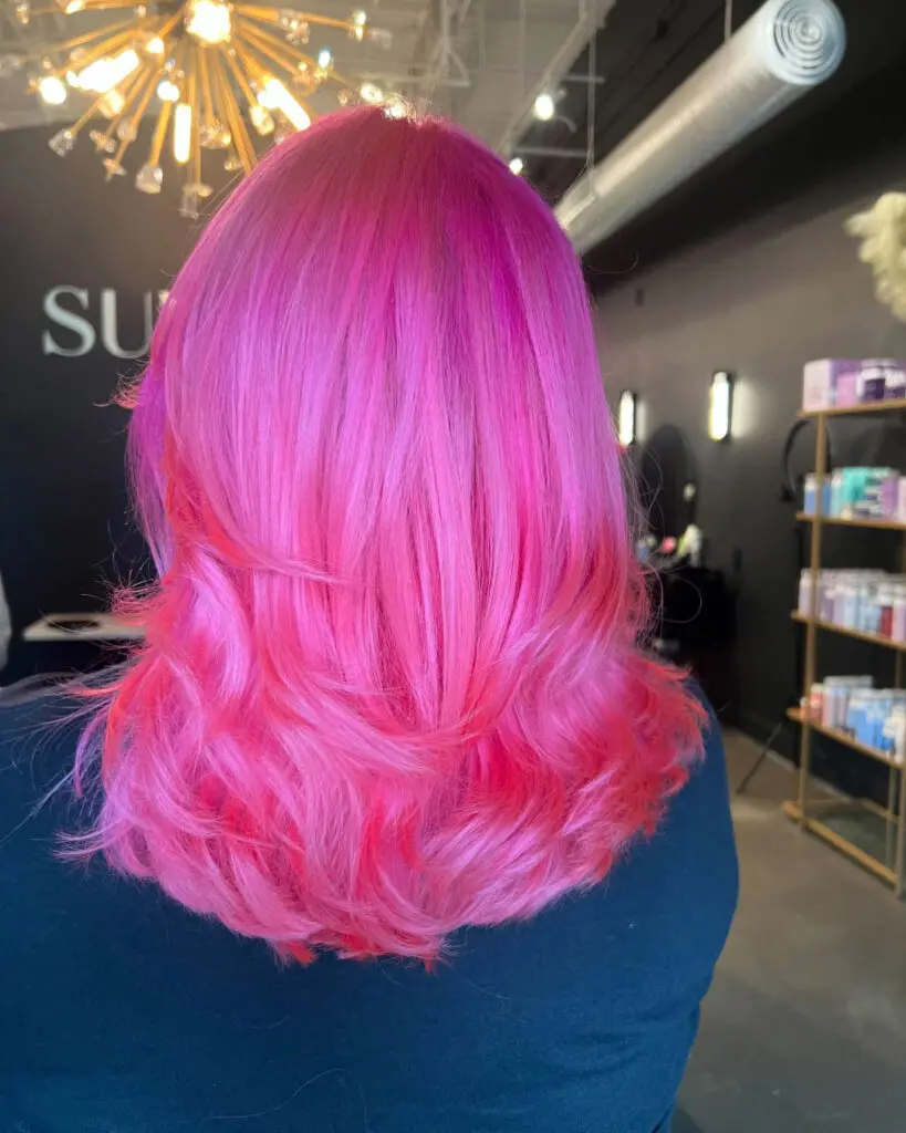 Back view of a client showcasing a vibrant, highly saturated neon pink hair color styled with bouncy, voluminous layered ends at Sunday Salon in Raleigh, NC.