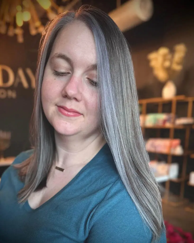 Woman with eyes closed showcasing long, sleek straight gray hair featuring a seamless silver color melt and a bright face-framing money piece, custom colored by Lisa Edwards at Sunday Salon in Raleigh, NC.