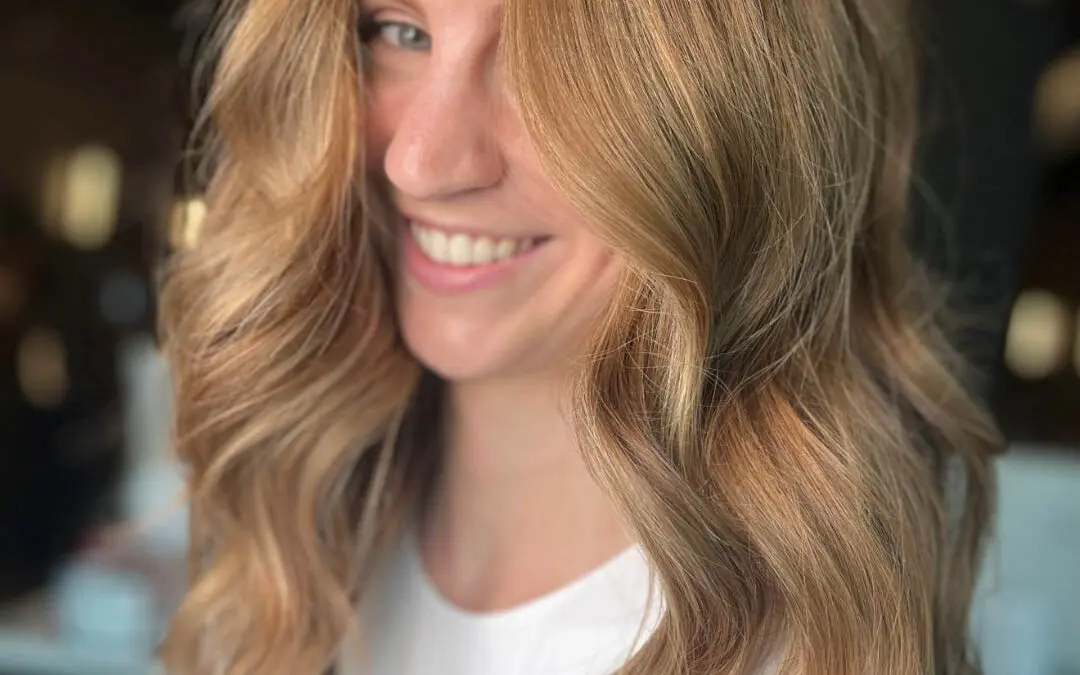 Smiling woman showcasing long, flowing hair with a dimensional golden copper and strawberry blonde balayage, styled with sweeping face-framing layers at Sunday Salon in Raleigh, NC.