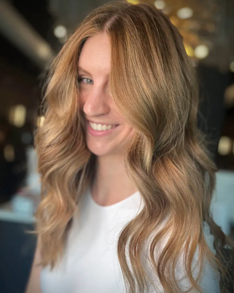 Radiant Golden Copper Balayage with Soft Cascading Layers Smiling woman showcasing long, flowing hair with a dimensional golden copper and strawberry blonde balayage, styled with sweeping face-framing layers at Sunday Salon in Raleigh, NC.
