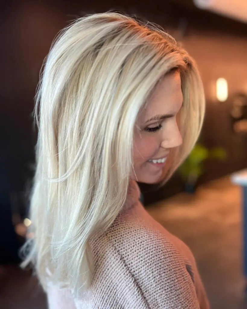 Luminous Creamy Vanilla Blonde with a Sleek Layered Blowout Side profile of a smiling woman with medium-long, bright creamy vanilla blonde hair styled in a sleek blowout with soft face-framing layers, cut and colored by Katherine Beingolea at Sunday Salon in Raleigh, NC.