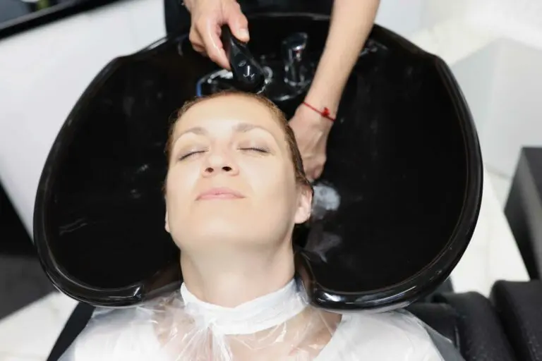 A person with closed eyes is reclining in a salon chair while getting their hair washed in a black sink by a hairstylist. Plastic protective coverings are draped around the person's neck and shoulders. The setting appears to be the best hair salon Raleigh, NC has to offer at Raleigh Iron Works.