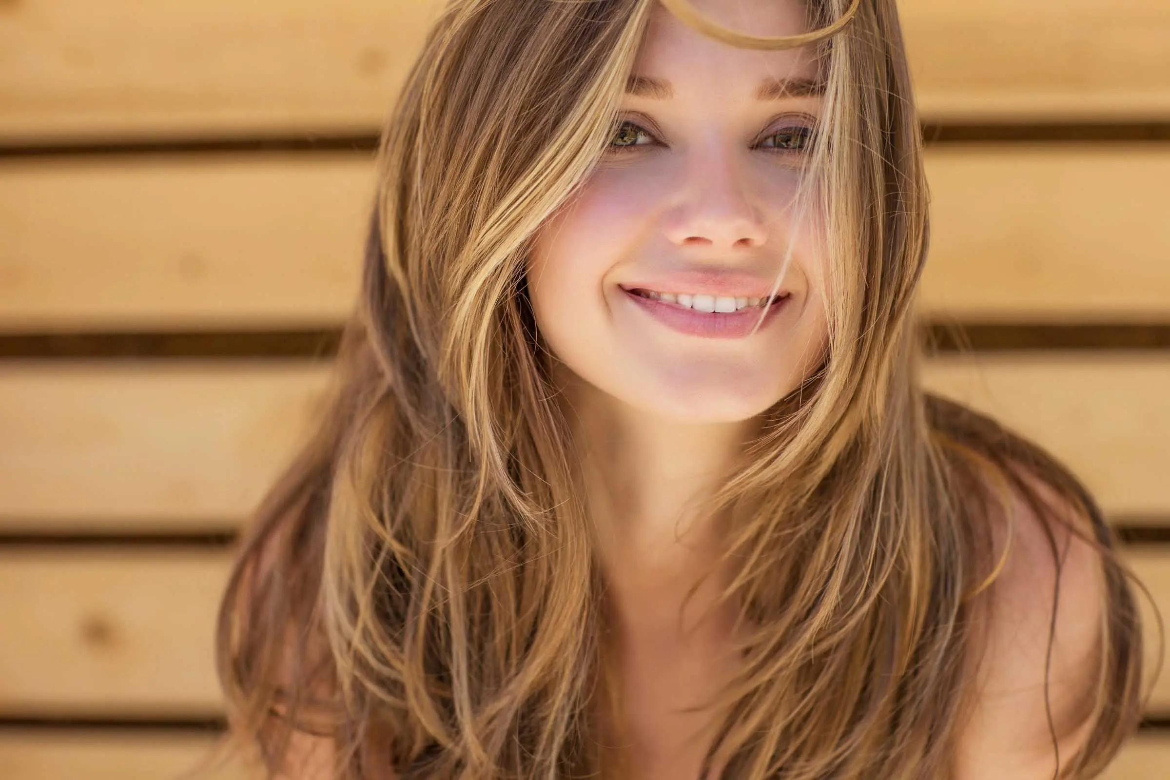 A woman with long, wavy blonde hair smiles warmly at the camera. Positioned in front of a background made of light wooden planks, she exudes a radiant glow. Her hair partially covers one of her eyes. For stunning styles like hers, visit one of the top hair salons Cary NC has to offer.