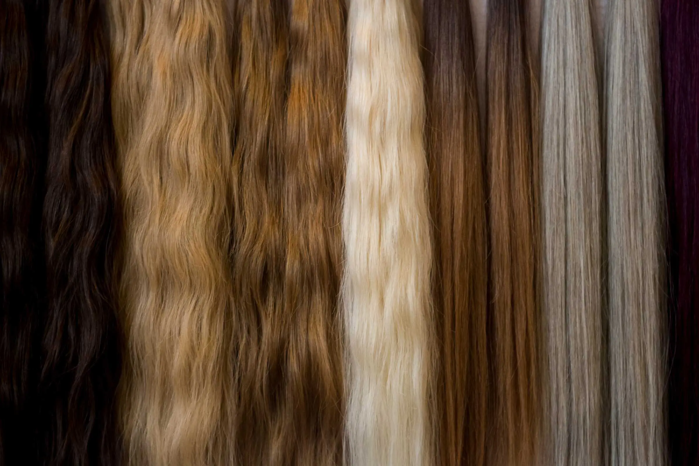 Close-up image of seven hair extensions, each in a different color and texture, ranging from dark brown, light brown, blonde, and white to shades of gray. The hair textures vary from wavy to straight. Perfect choices curated by the best hair salon Cary NC has to offer!
