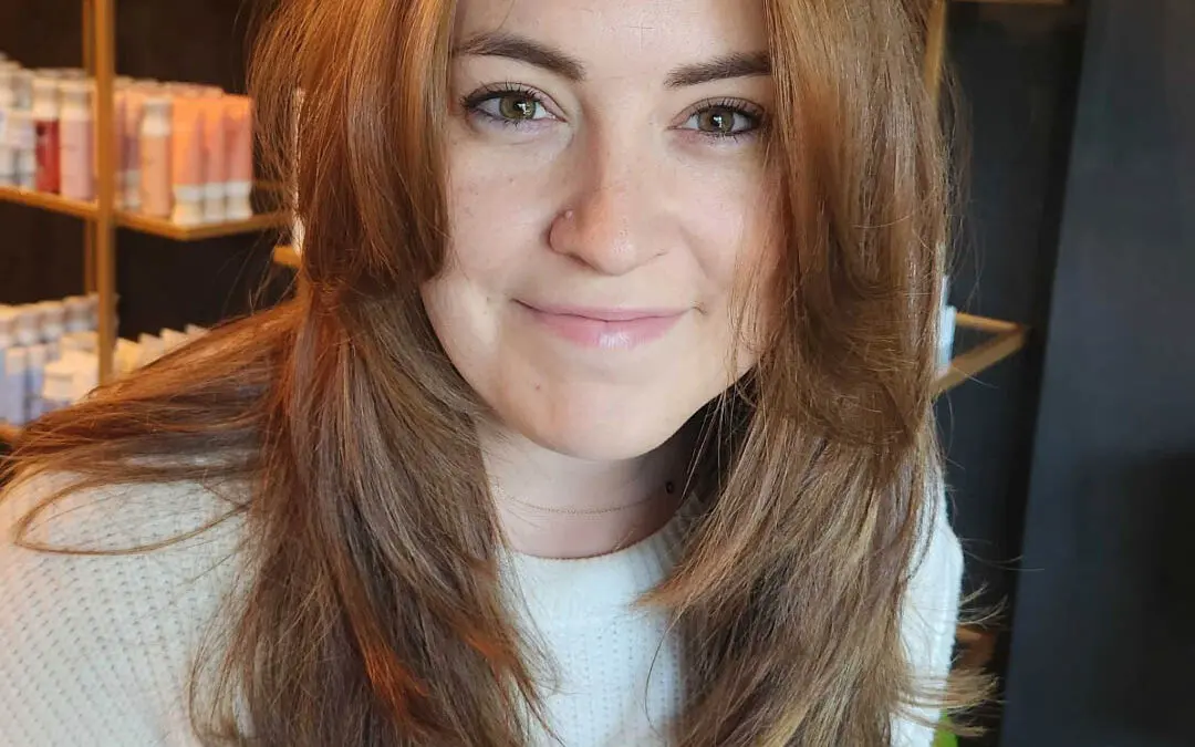 Front view of a smiling client with medium-long warm copper auburn hair, featuring sweeping curtain bangs and soft face-framing layers styled at Sunday Salon in Raleigh.