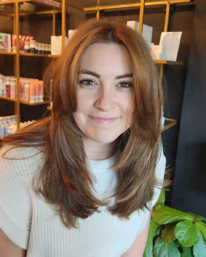 Warm Copper Auburn with Face-Framing Layers on Medium-Long Hair Front view of a smiling client with medium-long warm copper auburn hair, featuring sweeping curtain bangs and soft face-framing layers styled at Sunday Salon in Raleigh.