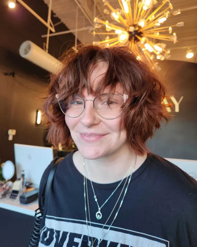 A smiling client at Sunday Salon in Raleigh wearing glasses, showcasing a vibrant auburn shag haircut with heavily textured waves and a piece-y fringe.