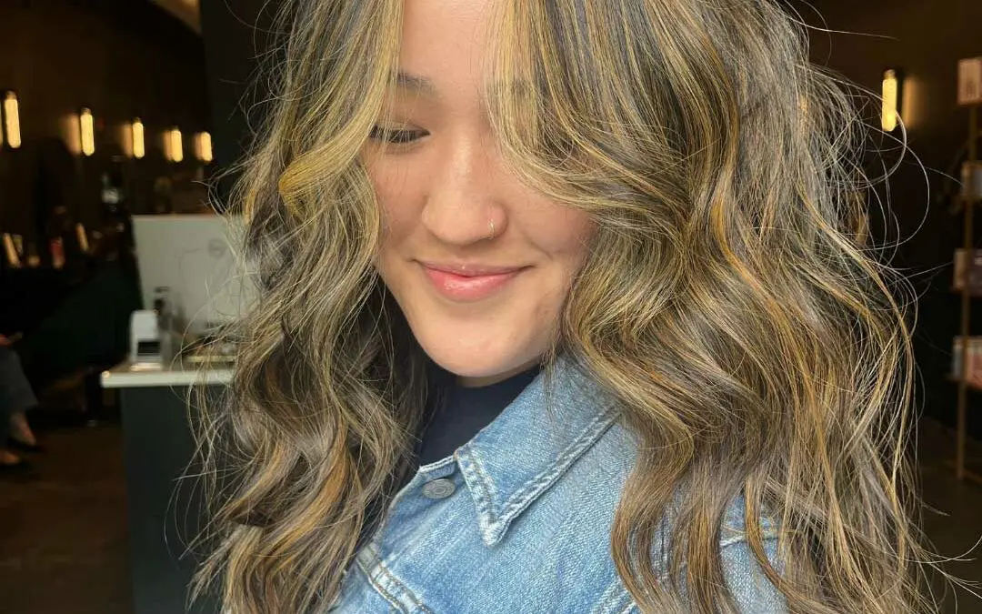 Front-facing view of a smiling client featuring long wavy brunette hair with warm golden caramel balayage and face-framing highlights at Sunday Salon in Raleigh.