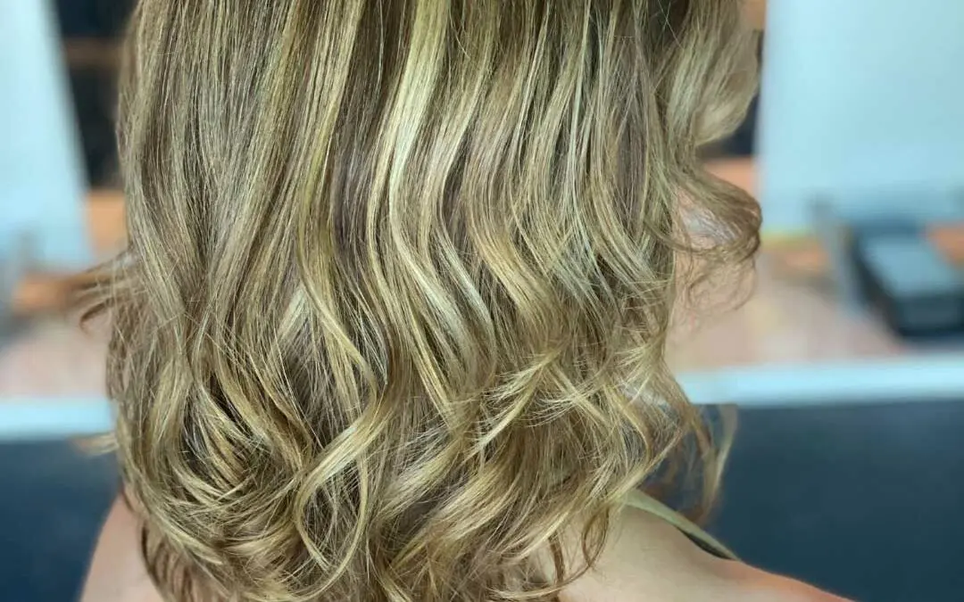 Back view of a client with shoulder-length layered hair featuring dimensional golden bronde highlights and bouncy curled ends at Sunday Salon in Raleigh.
