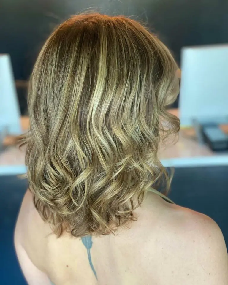 Back view of a client with shoulder-length layered hair featuring dimensional golden bronde highlights and bouncy curled ends at Sunday Salon in Raleigh.