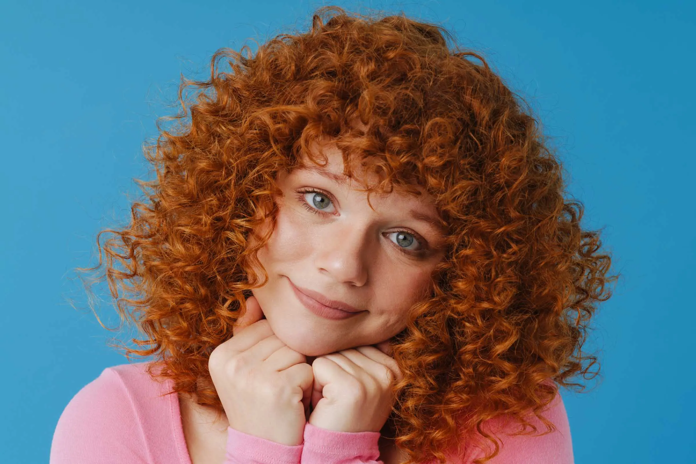 A person with curly red hair is smiling softly, resting their chin on their hands. They are wearing a pink shirt and are set against a blue background.