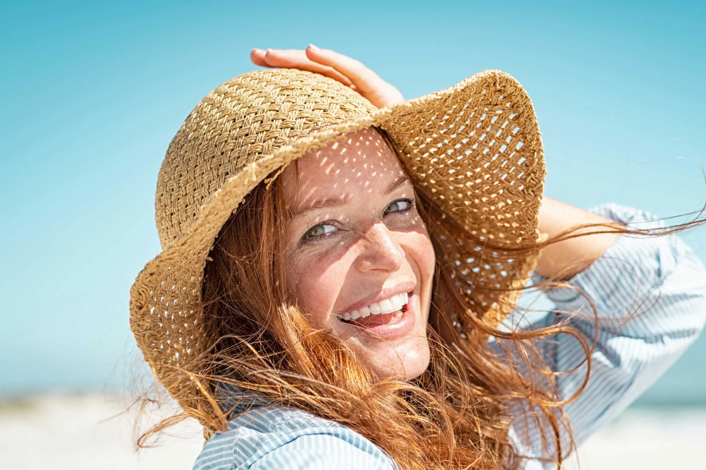 A woman with long, summer hair, wearing a straw hat, smiles brightly under a clear blue sky. The beach is blurred in the background, suggesting a sunny day. She holds the brim of her hat with one hand as her hair flows gently in the breeze—hair stylists' tips brought to life.