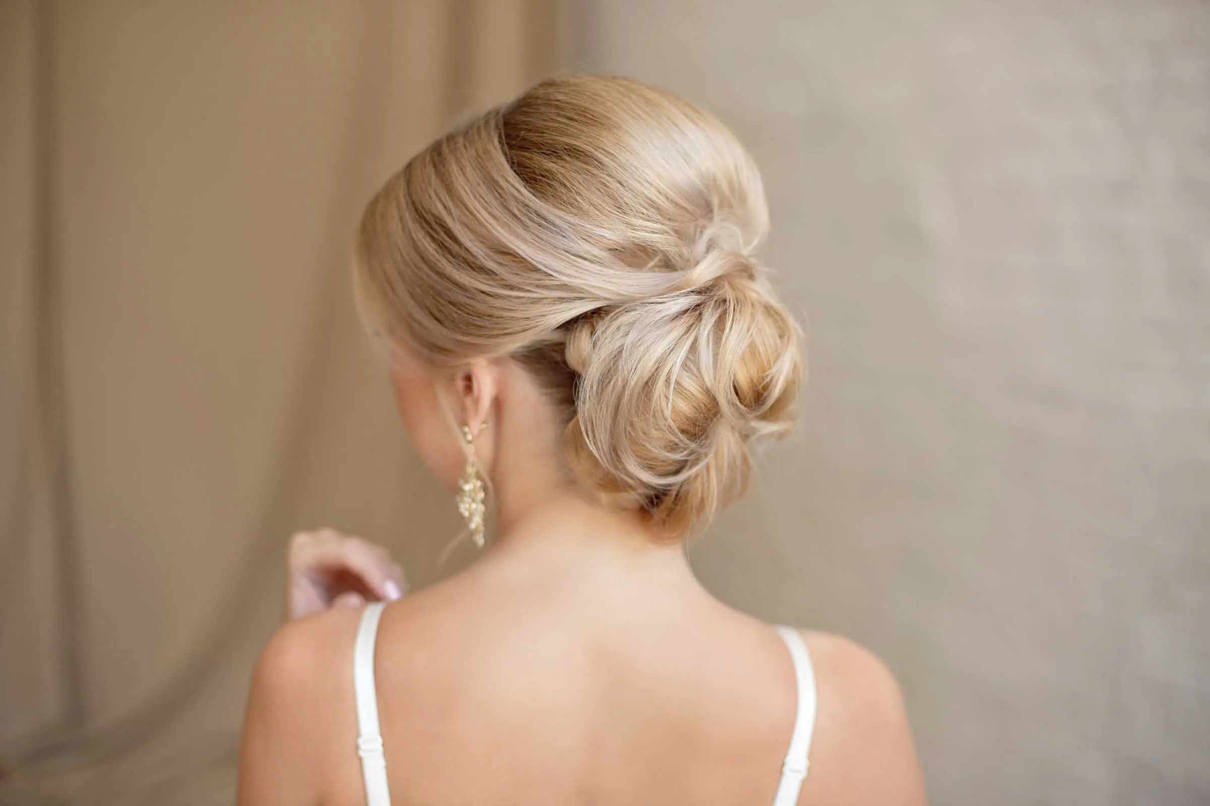 A woman with blonde hair styled in an elegant updo, featuring soft waves and a loose bun, is shown from behind. She is wearing a white spaghetti strap top and gold dangling earrings. The background is softly blurred with neutral tones, showcasing the expertise of formal hair styling services in Cary, NC.