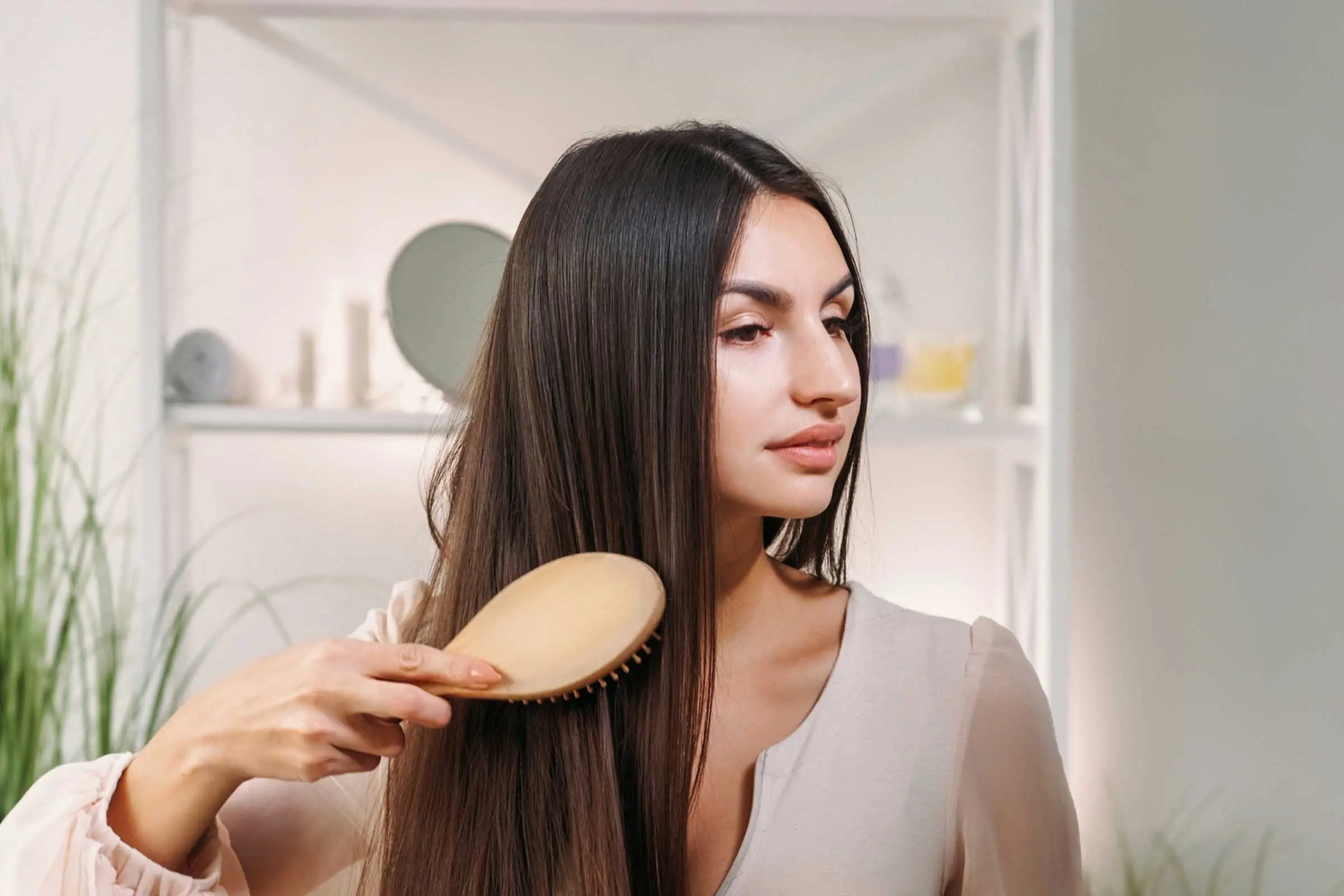 A woman with long, straight hair is brushing her hair with a wooden hairbrush. Indoors in a well-lit room with white walls and some plants in the background, she wears a light-colored blouse and looks slightly to her right. It's almost as if the scene was designed by Auto Draft for its serene simplicity.