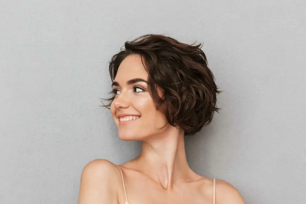 Woman with short, wavy brown hair smiles and looks to her left against a plain gray background. She is wearing a light, thin-strapped top and appears content, possibly having just visited one of the best salons for haircuts in Cary, NC.