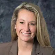 A person with long blonde hair is smiling at the camera. They are wearing a brown blazer, and the background is a textured dark gray.