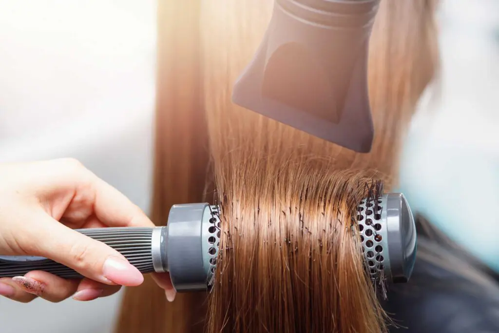 Close-up of a person blow-drying their hair for a sleek blowout. A round brush is used to smooth and style the long, straight, brown hair. A blow-dryer with a concentrator nozzle attachment directs hot air onto the hair being rolled around the brush.