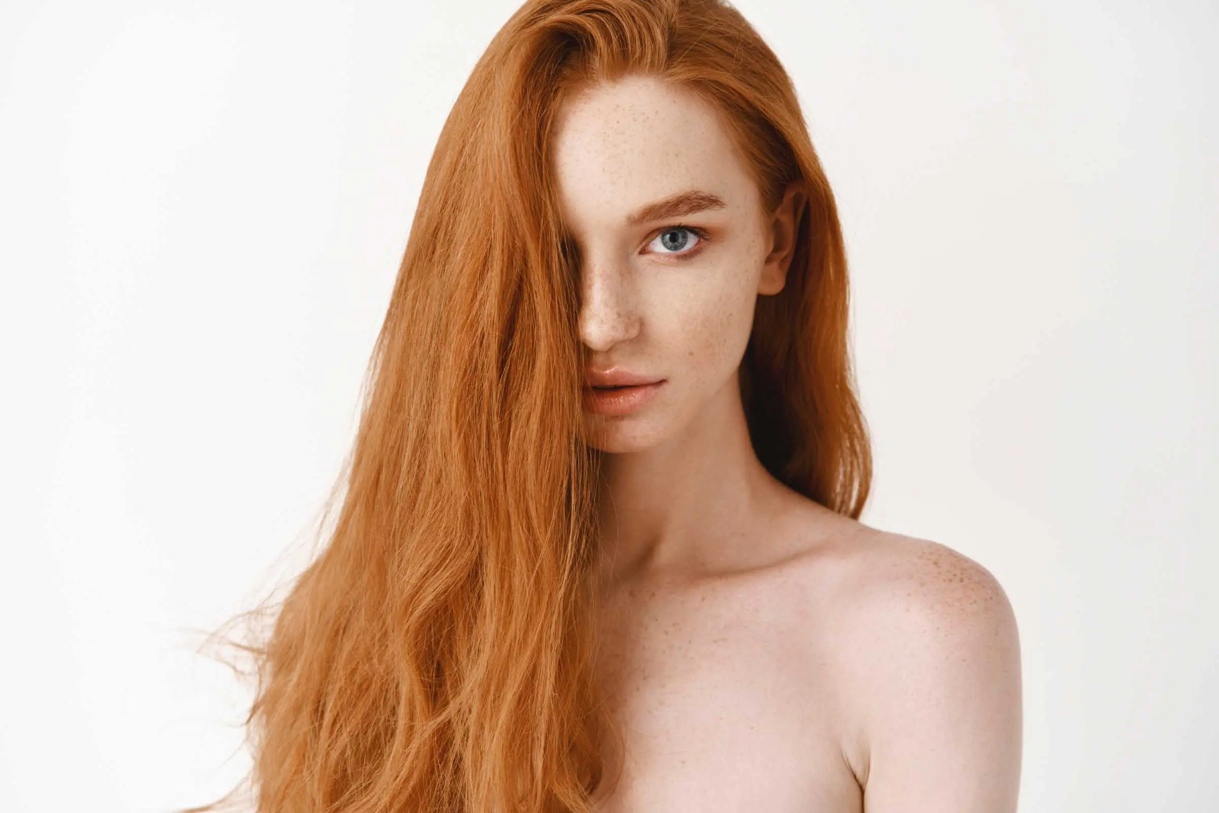 A person with long, red hair, showcasing an all-over color, partially covering one eye gazes directly at the camera. They have fair skin with freckles and are set against a plain white background. The image captures a minimalistic and natural aesthetic.
