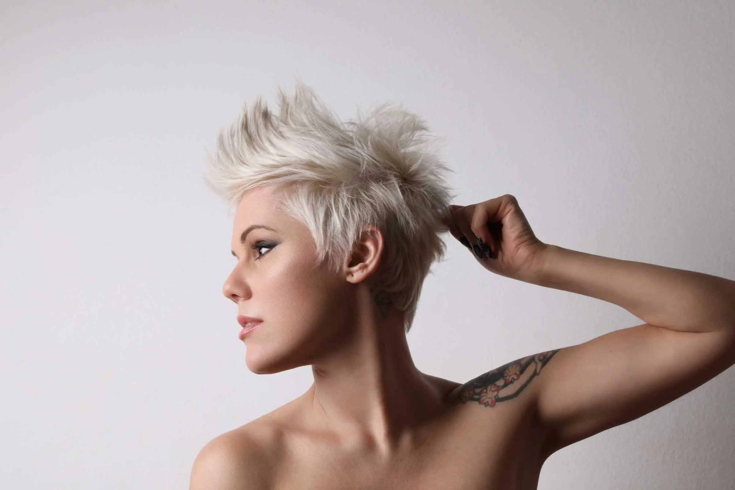 A person with a professional haircut—a short, platinum blonde mohawk—is looking to the side, touching their hair with one hand. The individual has a visible tattoo on their upper arm. The background is plain white.