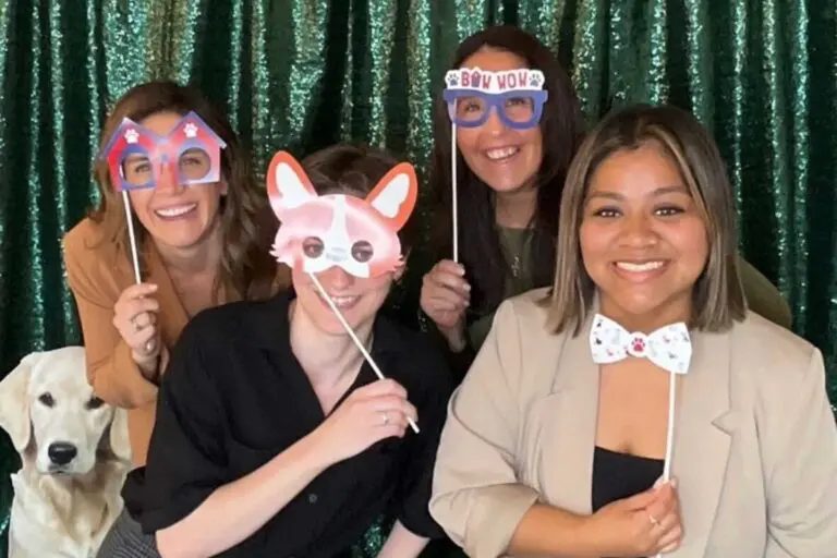Four people pose playfully with novelty glasses and props in front of a green glittery backdrop at the Neuse River Golden Retriever Rescue Gala. A golden retriever stands beside them, adding charm to the lively scene celebrating sponsorship and support.