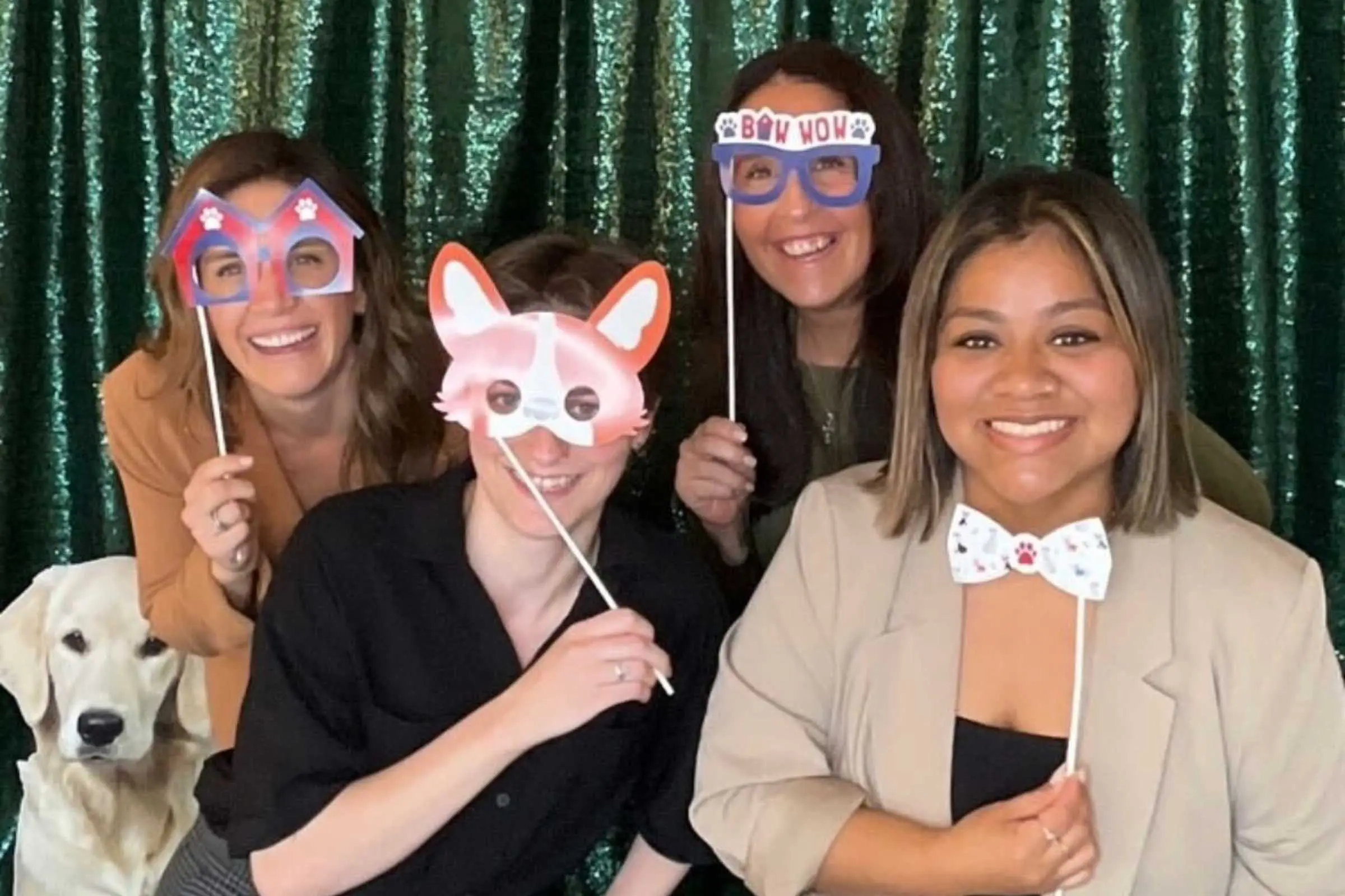 Four people pose playfully with novelty glasses and props in front of a green glittery backdrop at the Neuse River Golden Retriever Rescue Gala. A golden retriever stands beside them, adding charm to the lively scene celebrating sponsorship and support.