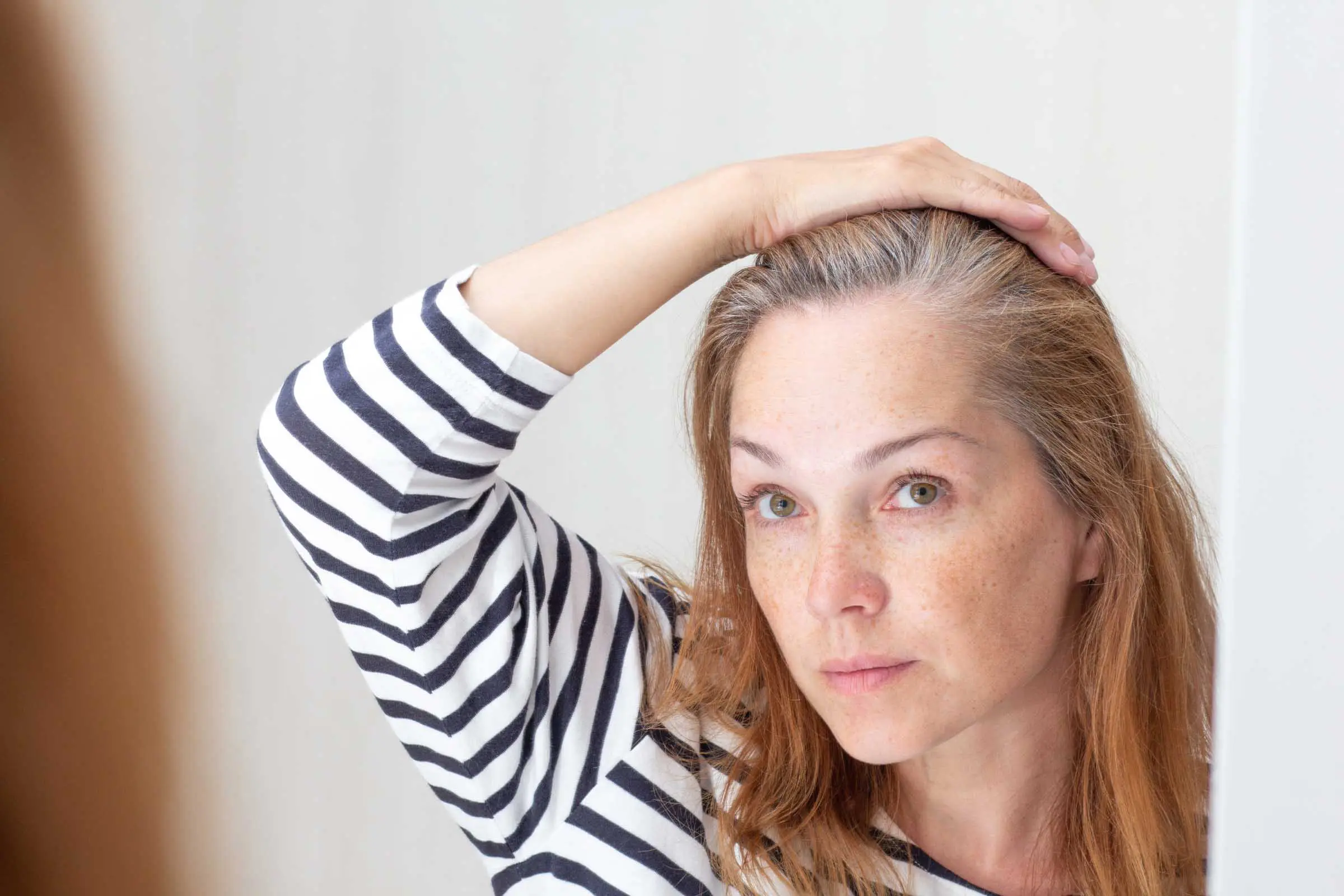 A person with freckles and long hair, wearing a striped shirt, looks into a mirror and holds their hair back with one hand to reveal gray roots, contemplating grey coverage. The background is a light, neutral color.