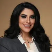 Smiling woman with long dark hair, wearing a gray blazer and white blouse, against a beige background.