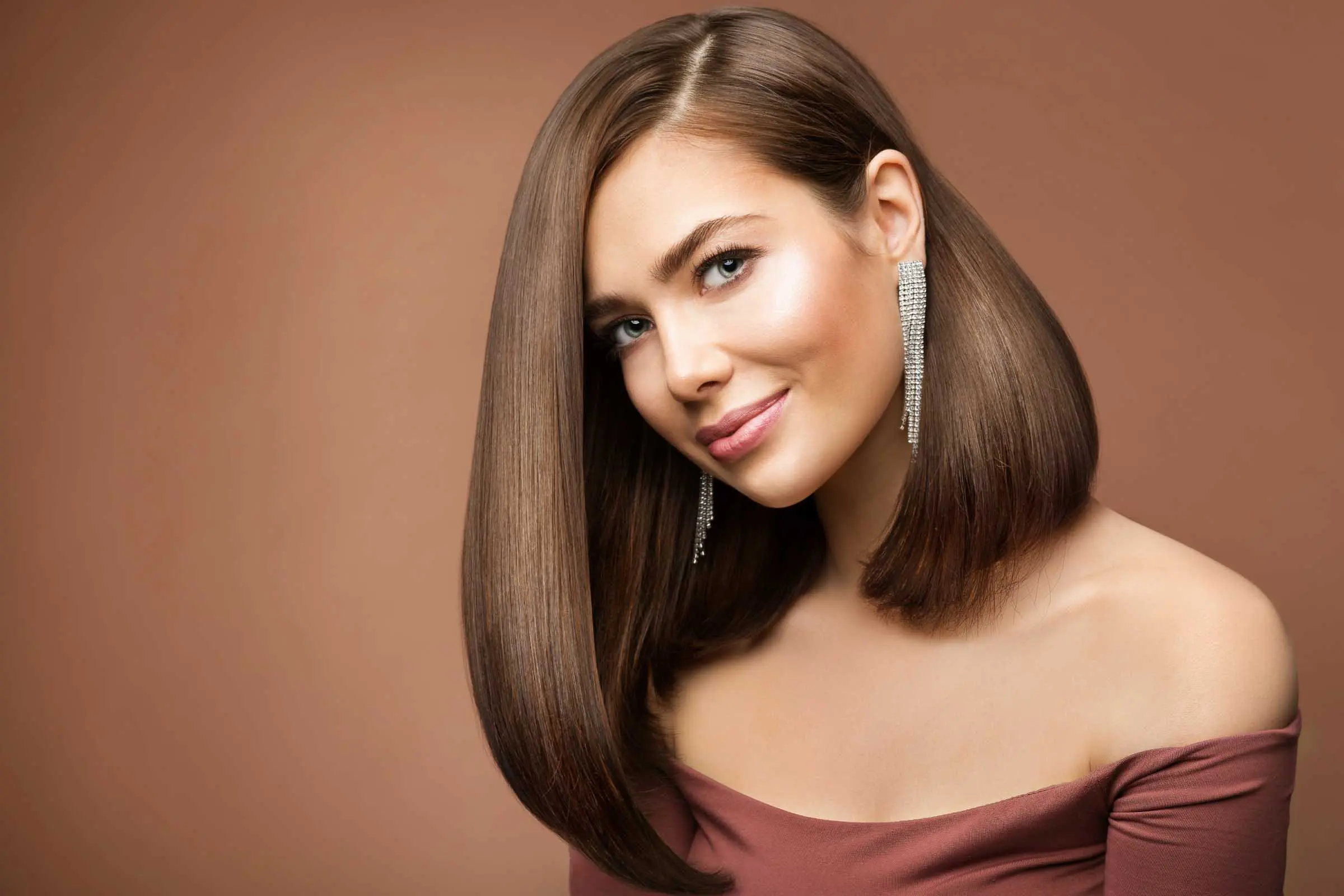 A woman with the perfect haircut, featuring straight, shoulder-length brown hair, smiles at the camera. She wears long, sparkling earrings and an off-the-shoulder brown top. The warm, light brown background beautifully complements her attire and hair.